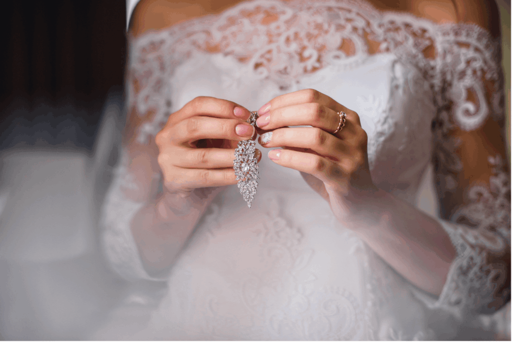 Eine Braut in einem wunderschönen weißen Brautkleid mit Spitzenbesatz im Off-Shoulder-Stil hält einen funkelnden, opulenten Kristallohrring in ihren Händen.