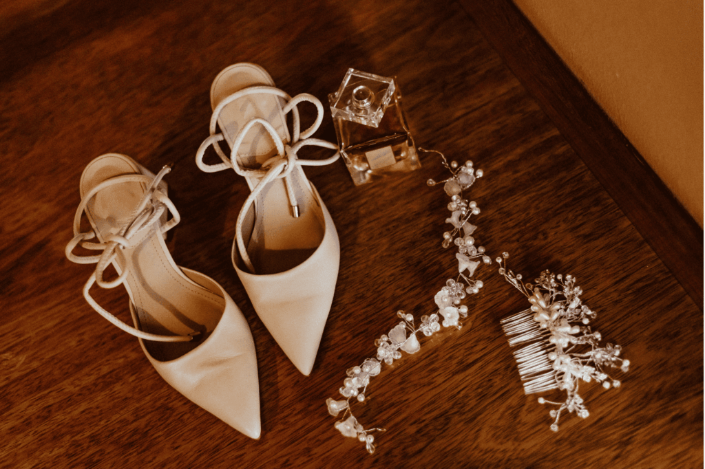 Eine elegante Zusammenstellung von Hochzeitsaccessoires auf einem dunklen Holzboden: Zwei cremefarbene Brautschuhe mit Riemchenschnürung liegen neben einem Flakon Parfum und filigranem Haarschmuck.