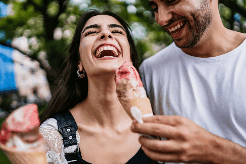 Ein glückliches Paar lacht und isst Eis im Sommer. Die Frau hält ein rosafarbenes und braunes Eis, während der Mann neben ihr steht und lächelt.