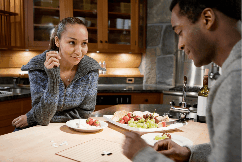 Ein lächelndes Paar spielt ein Brettspiel an einem Holztisch in einer modern eingerichteten Küche, auf dem auch eine Obstplatte mit Erdbeeren und Trauben steht.