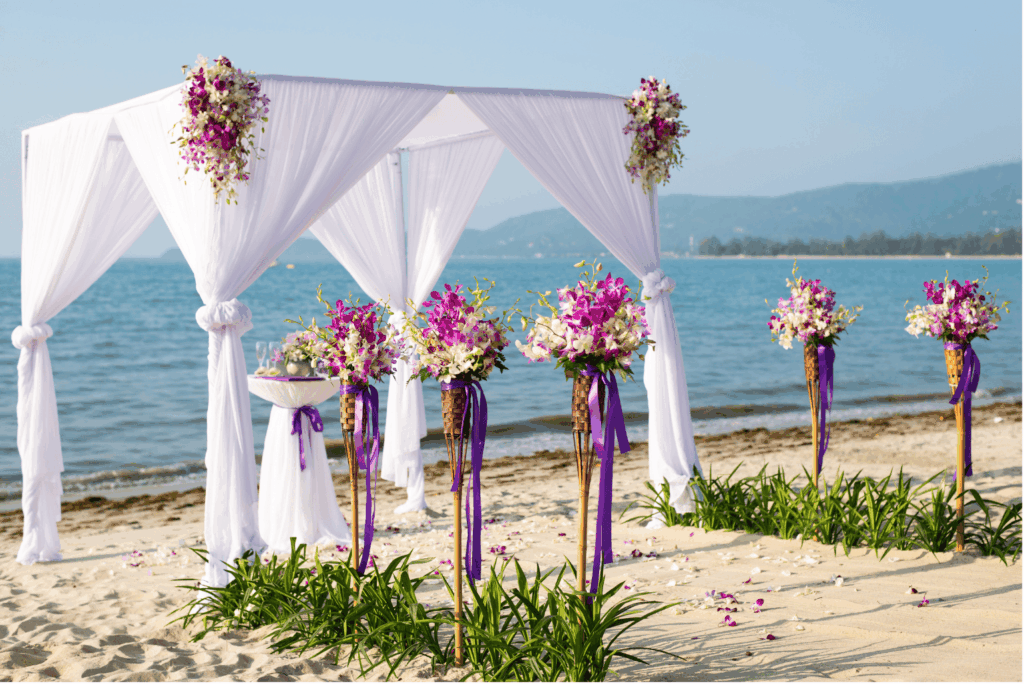 Ein wunderschön dekorierter Hochzeits-Pavillon am Strand, geschmückt mit weißen Stoffen und lila-pinken Blumenarrangements, bereit für eine romantische Zeremonie mit dem Meer und Bergen im Hintergrund.
