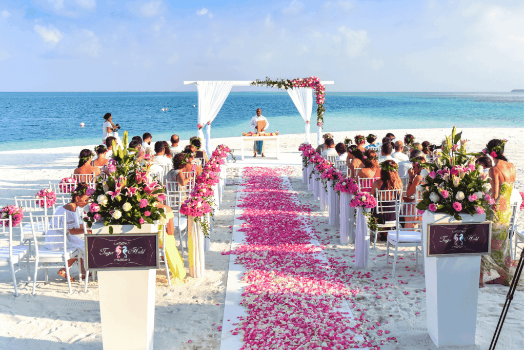 Eine romantische Strandhochzeit bei strahlendem Sonnenschein, mit einem mit Rosenblättern übersäten Gang, der zu einem geschmückten Hochzeitsbogen führt, und Gästen, die auf weißen Stühlen sitzen, während das türkisfarbene Meer im Hintergrund glitzert.
