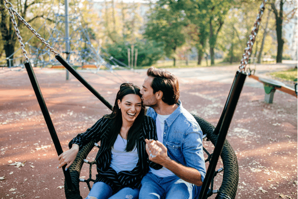Ein glückliches Paar sitzt auf einer Schaukel in einem Park. Der Mann küsst die Frau auf die Stirn, und sie lacht mit geschlossenen Augen. Sie halten Händchen und wirken sehr verliebt. Im Hintergrund sind Bäume und ein Spielplatz zu sehen.