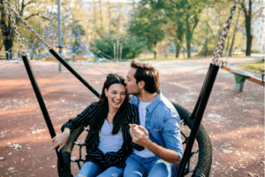 Ein glückliches Paar sitzt auf einer Schaukel in einem Park. Der Mann küsst die Frau auf die Stirn, und sie lacht mit geschlossenen Augen. Sie halten Händchen und wirken sehr verliebt. Im Hintergrund sind Bäume und ein Spielplatz zu sehen.