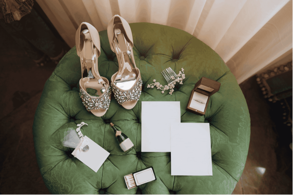 Elegante Brautschuhe mit Strassbesatz auf einem grünen Samthocker, daneben ein Verlobungsring, eine kleine Sektflasche und leere Karten für einen emotionalen Moment.