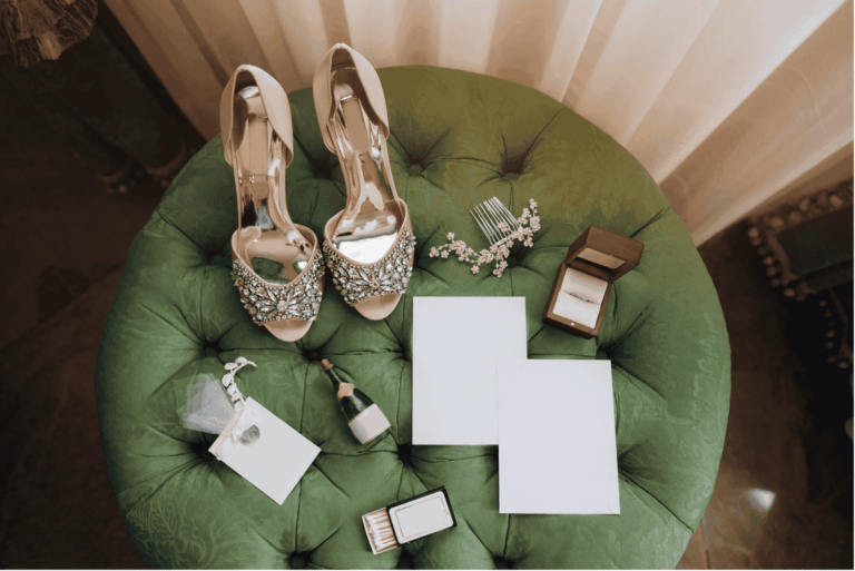 Elegante Brautschuhe mit Strassbesatz auf einem grünen Samthocker, daneben ein Verlobungsring, eine kleine Sektflasche und leere Karten für einen emotionalen Moment.