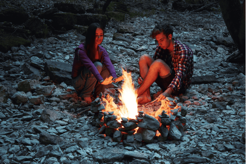 Ein junges Paar sitzt barfuß und nah beieinander an einem knisternden Lagerfeuer, das von einem Kreis aus Steinen umgeben ist, in einer dunklen, felsigen Umgebung. Sie schauen beide auf die Flammen, die ihre Gesichter in warmes Licht tauchen.