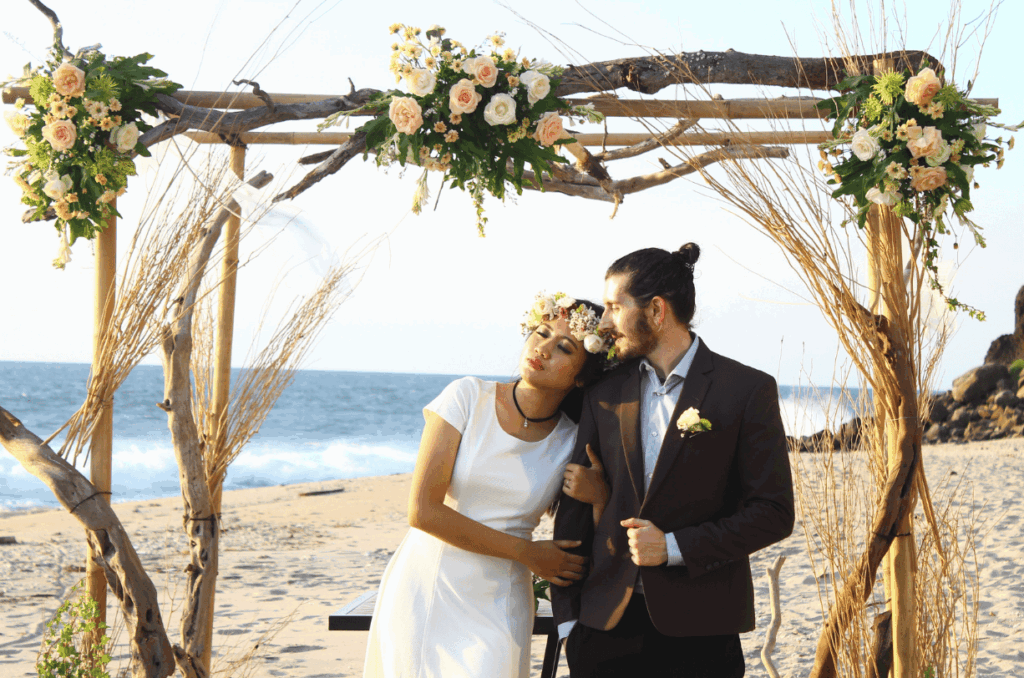Braut und Bräutigam unter einem mit Blumen geschmückten Traubogen am Strand bei Sonnenuntergang