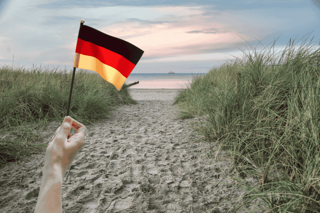 Eine Hand hält eine kleine deutsche Flagge auf einem Sandweg, der zu einem Strand mit Meer und bewachsenen Dünen führt. Der Himmel ist bewölkt mit einem Sonnenuntergang.