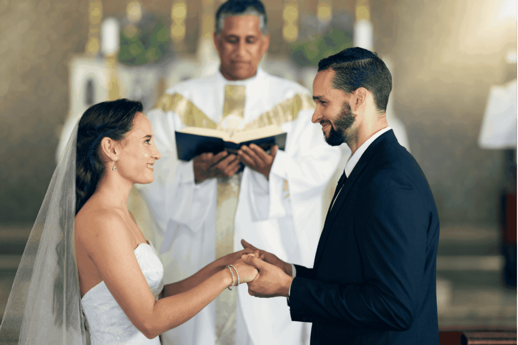 Ein Paar steht sich während seiner Hochzeitszeremonie gegenüber, hält Händchen und lächelt. Ein Priester in einem weißen Gewand hält im Hintergrund ein Buch in den Händen.