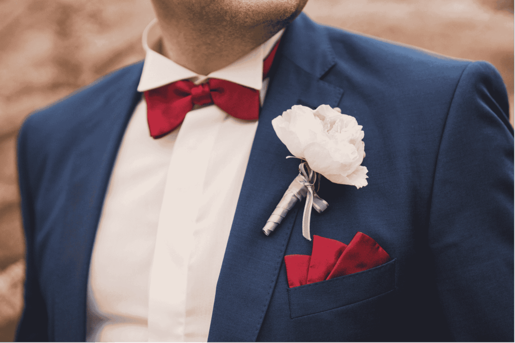 Ein Mann trägt einen dunkelblauen Anzug mit einer roten Fliege, einem weißen Hemd, einer weißen Boutonniere und einer roten Einstecktuch.