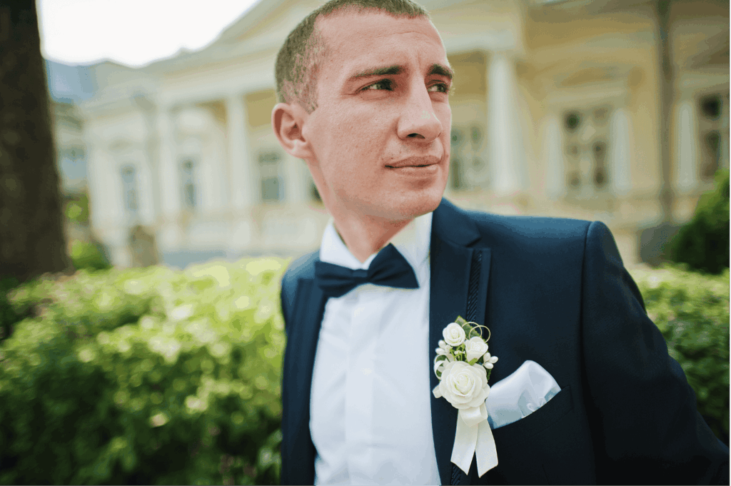 Ein Bräutigam im dunkelblauen Anzug mit weißem Hemd, Fliege und Boutonnière schaut nachdenklich zur Seite, mit einem eleganten Gebäude im Hintergrund.