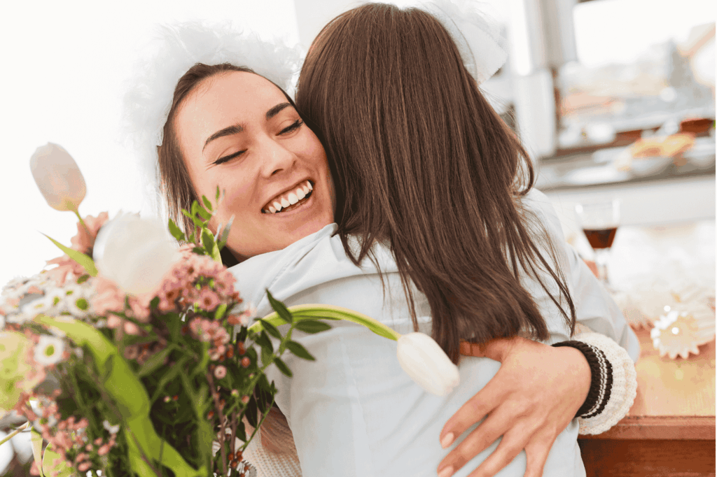 Eine Frau mit einem Schleier auf dem Kopf und einem Blumenstrauß in der Hand wird von einer anderen Frau herzlich umarmt. Beide lächeln vor Freude.