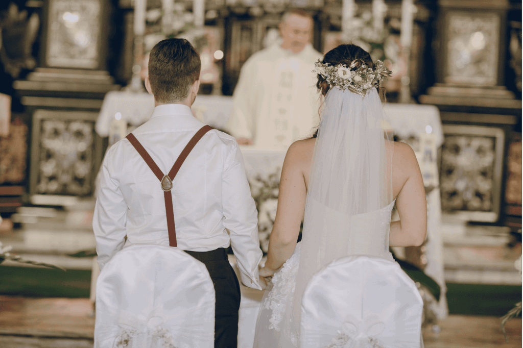 Braut und Bräutigam sitzen mit dem Rücken zur Kamera während einer Hochzeitszeremonie in einer Kirche, sie halten Händchen. Im Hintergrund steht ein Priester.