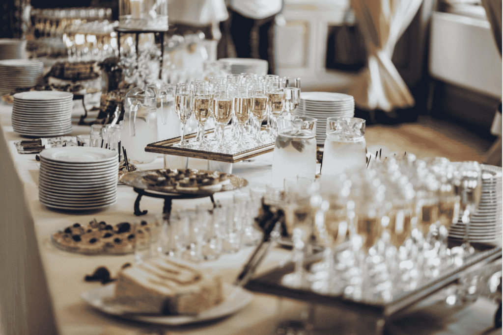 Ein reich gedecktes Buffet mit gestapelten Tellern, Kuchenstücken, kleinen Snacks, Krugwasser und mehreren Reihen gefüllter Sektgläser auf Tabletts, bereit für die Gäste.