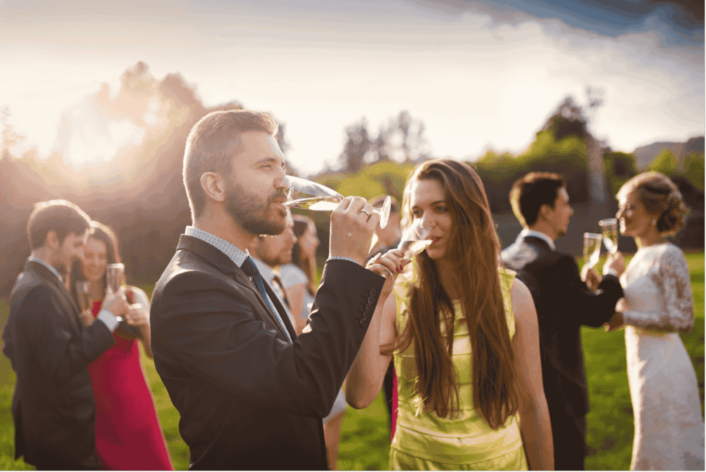 Ein Mann und eine Frau in festlicher Kleidung stoßen bei einem Sektempfang im Freien an. Die Sonne scheint warm im Hintergrund. Die Gäste genießen den Aperitif auf einer Feier oder Hochzeit.