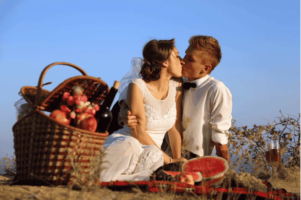 Brautpaar küsst sich innig bei einem romantischen Picknick im Freien, mit Picknickkorb, Wein und Wassermelone, vor einem weiten blauen Himmel.
