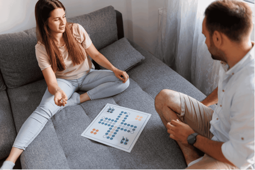 Ein lächelndes Paar sitzt auf einer grauen Couch und spielt gemeinsam ein einfaches Brettspiel (Mensch ärgere dich nicht ähnliches Layout).