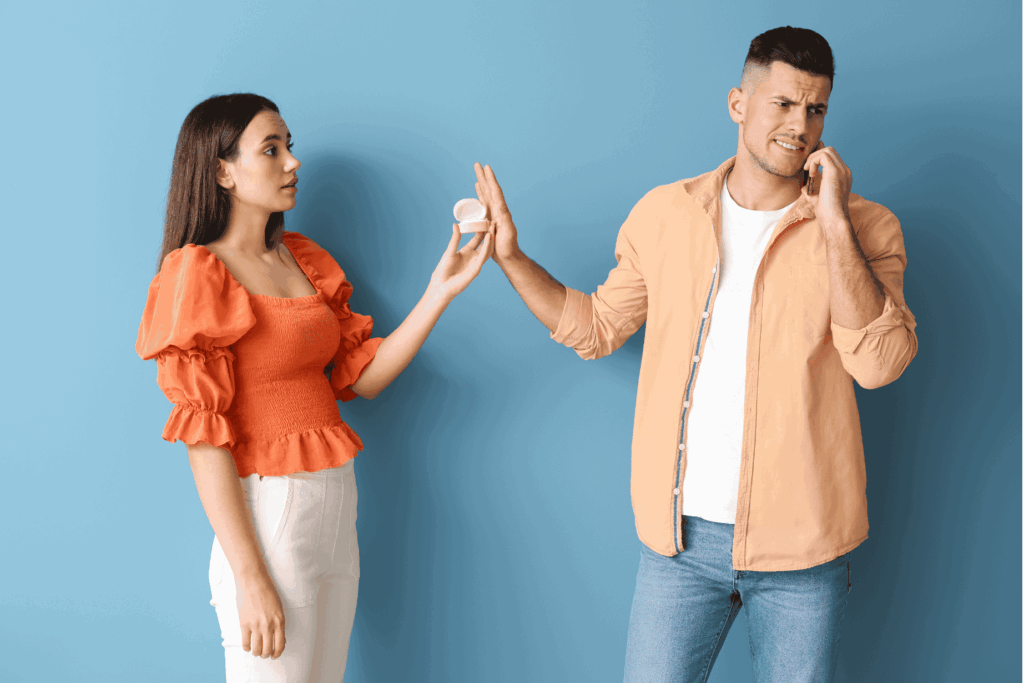 Eine Frau hält einem Mann eine offene Ringschachtel hin. Der Mann macht ein abwehrendes oder verwirrtes Gesicht, während er die Hand ablehnend entgegenstreckt. Der Hintergrund ist blau.