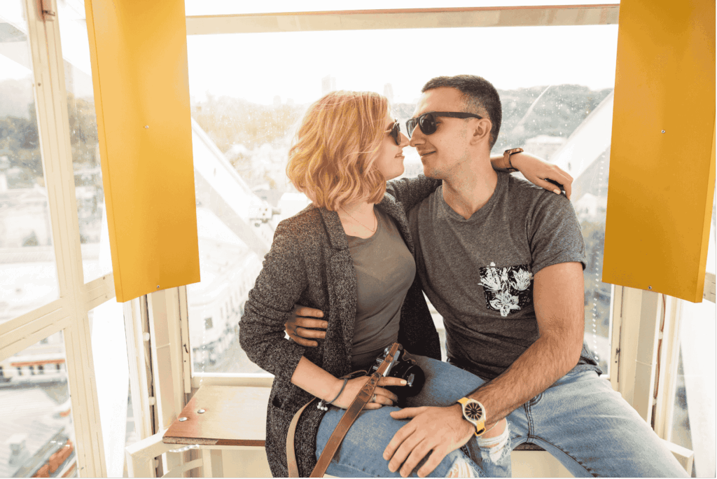 Eine Frau und ein Mann sitzen eng umschlungen in einer Gondel eines Riesenrads und schauen sich verliebt an. Die Frau trägt einen Ring an der Hand, der Mann hat seinen Arm um sie gelegt.