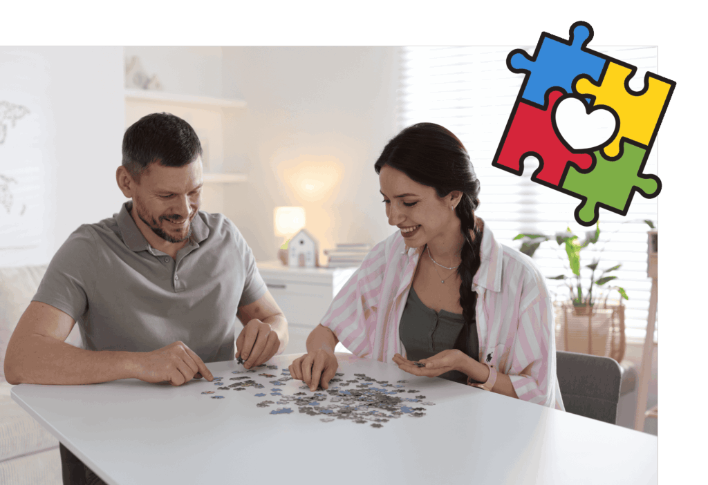 Ein Mann und eine Frau sitzen an einem Tisch und legen ein Puzzle. Die Frau lächelt. Rechts oben ist eine Grafik mit vier bunten Puzzleteilen, die in der Mitte ein Herz bilden, eingeblendet.