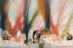 Ein Brautpaar küsst sich innig vor einem spektakulären Hintergrund aus einer Explosion von farbigem Rauch (Rot, Gelb, Grün, Blau). Vor ihnen steht die Hochzeitstorte.