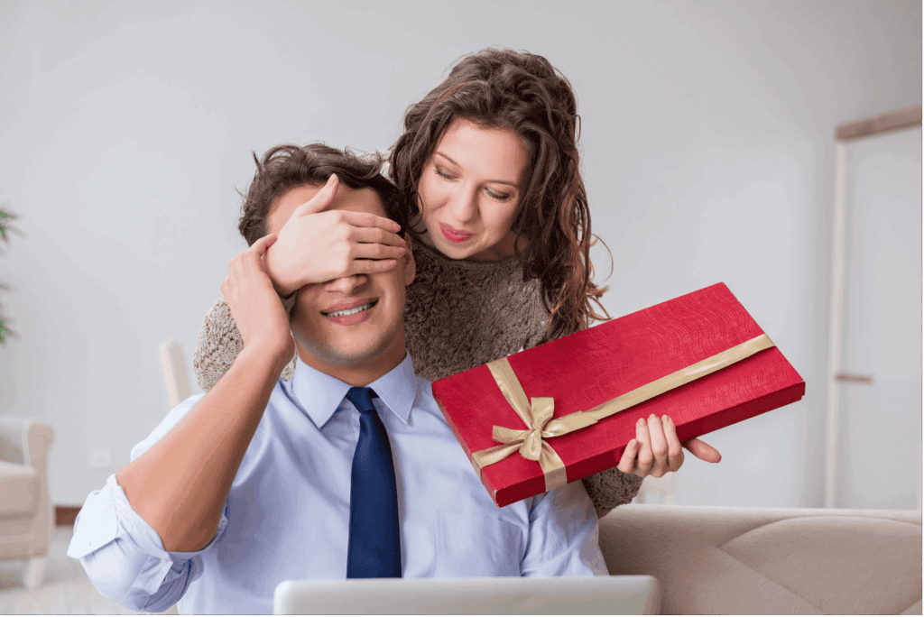 Eine lächelnde Frau hält einem sitzenden Mann von hinten die Augen zu und überreicht ihm eine flache, rechteckige, rot verpackte Geschenkschachtel mit goldener Schleife.