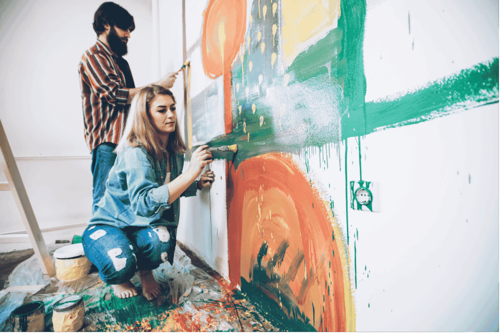 Ein Paar malt gemeinsam und konzentriert mit Pinsel und Farbe abstrakte, farbenfrohe Muster in Grün und Orange an eine weiße Wand.