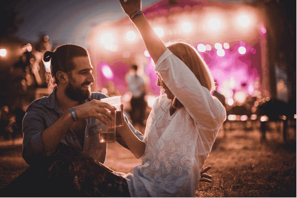 Ein glückliches Paar sitzt auf dem Gras eines Open-Air-Konzerts am Abend, hält Getränke in der Hand und schaut sich an, während die Bühne im Hintergrund hell leuchtet.