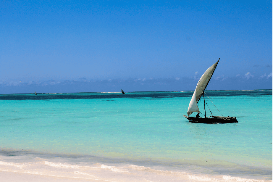 Ein traditionelles Segelboot (Dhau) segelt über das extrem türkisfarbene Wasser vor einem weißen Sandstrand, typisch für Sansibar.