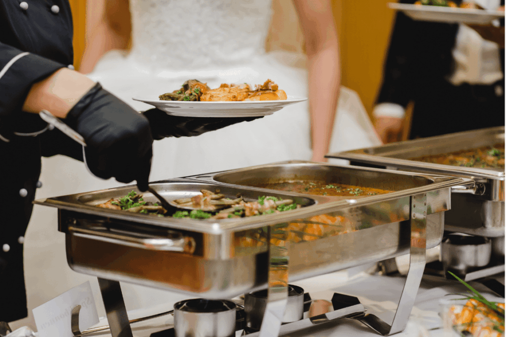 Ein Koch mit schwarzen Handschuhen serviert Speisen von einem Buffet auf einem Teller. Im Hintergrund ist undeutlich eine Braut in einem weißen Kleid zu sehen.