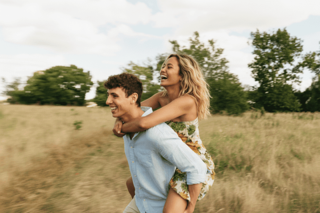 Ein lachendes Paar auf einer sommerlichen Wiese; ein Mann trägt eine Frau im Piggyback-Style (Huckepack), beide wirken ausgelassen und glücklich vor einem Hintergrund aus Bäumen und hellem Himmel.
