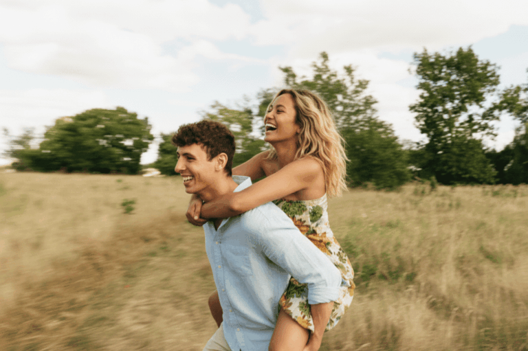 Ein lachendes Paar auf einer sommerlichen Wiese; ein Mann trägt eine Frau im Piggyback-Style (Huckepack), beide wirken ausgelassen und glücklich vor einem Hintergrund aus Bäumen und hellem Himmel.