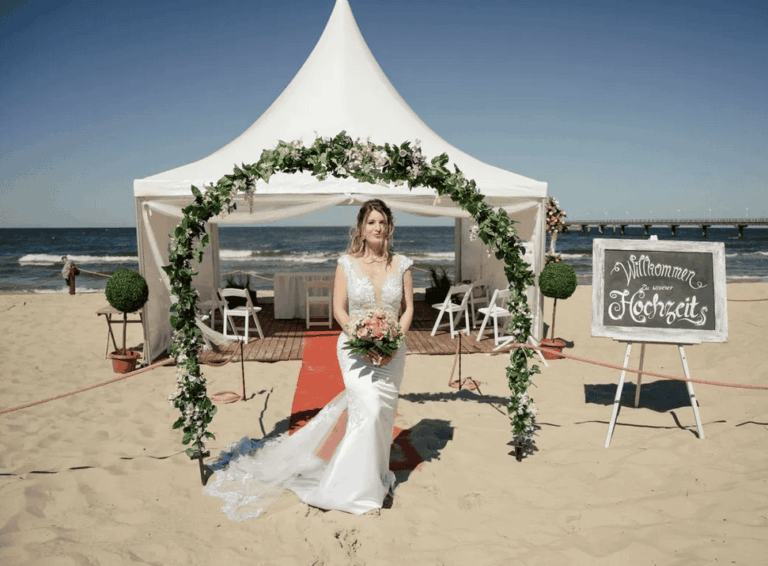 Eine Braut im eleganten Hochzeitskleid steht unter einem Blumenbogen am Strand von Zinnowitz auf Usedom. Im Hintergrund sind ein weißes Trauzelt, die Ostsee und die Seebrücke zu sehen.